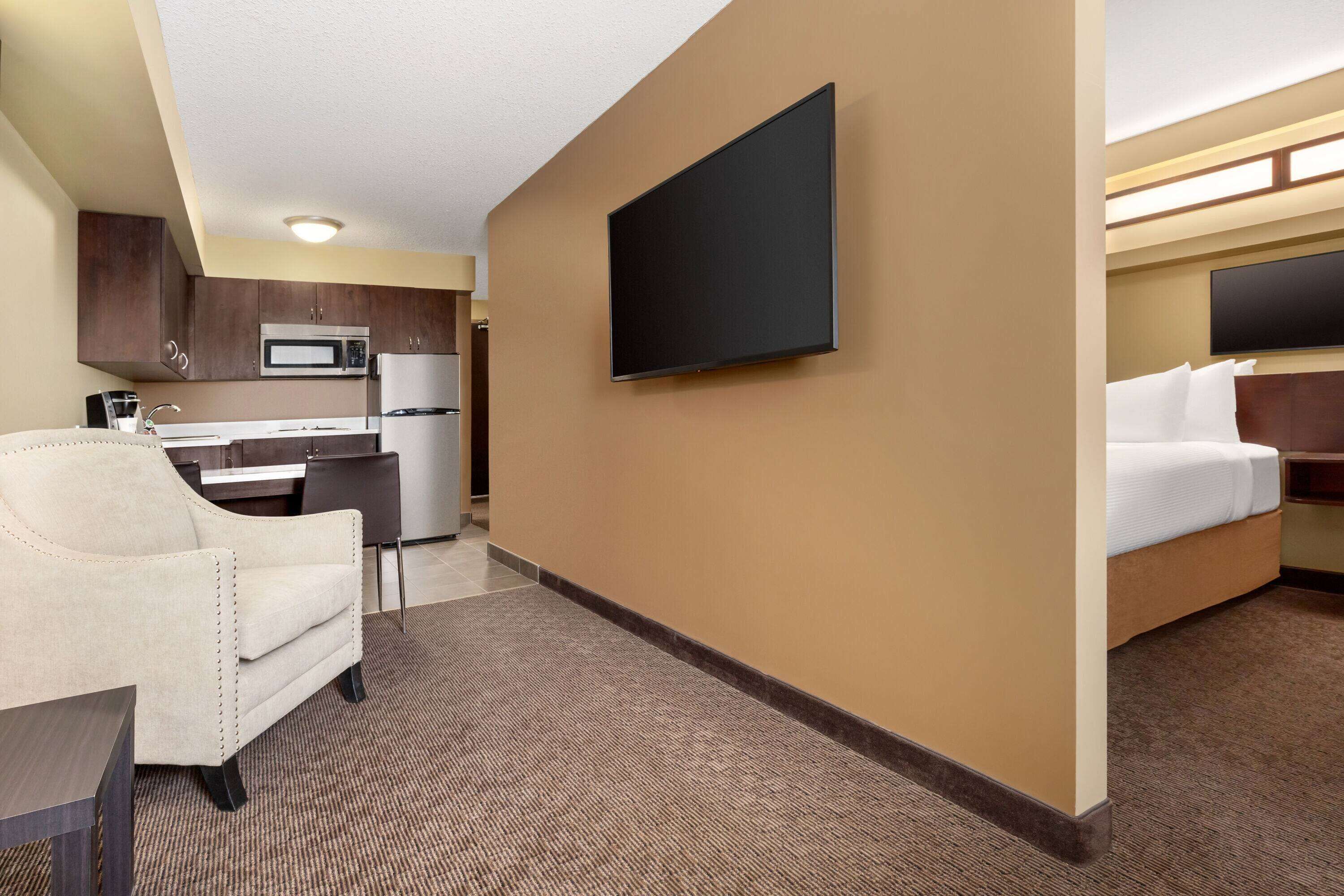 Ein Hotelzimmer mit einem Bett, einem an der Wand montierten Fernseher, einer Küchenzeile mit Mikrowelle und Kühlschrank sowie einer Sitzecke mit Sessel und Tisch.