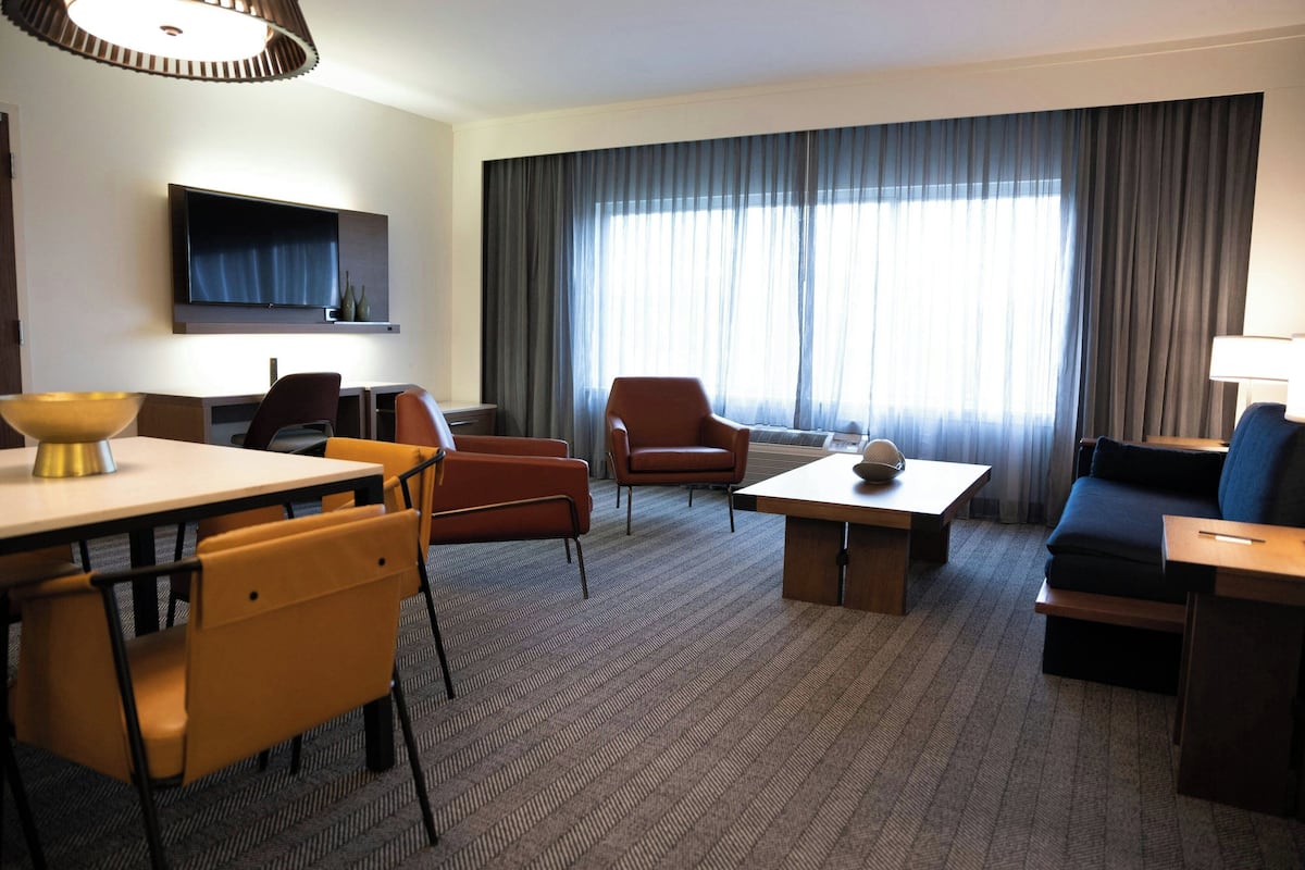 Hotellihuone, jossa on seinälle asennettu taulutelevisio, ruokailutila pöytineen ja tuoleineen, sohva sekä suuri ikkuna, jossa on verhot.