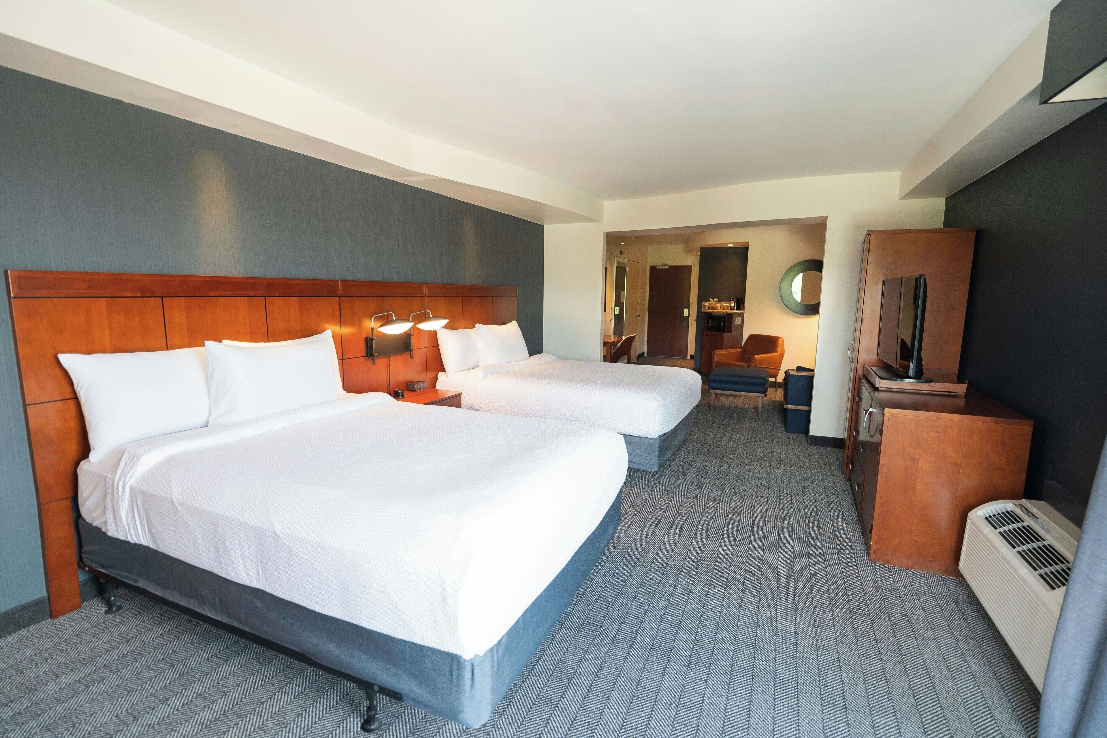 Une chambre d’hôtel avec deux lits, une tête de lit en bois, un bureau avec une chaise, une télévision et un miroir.