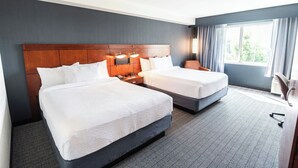 A hotel room with two beds, a wooden headboard, bedside lamps, a desk, and a window with curtains.