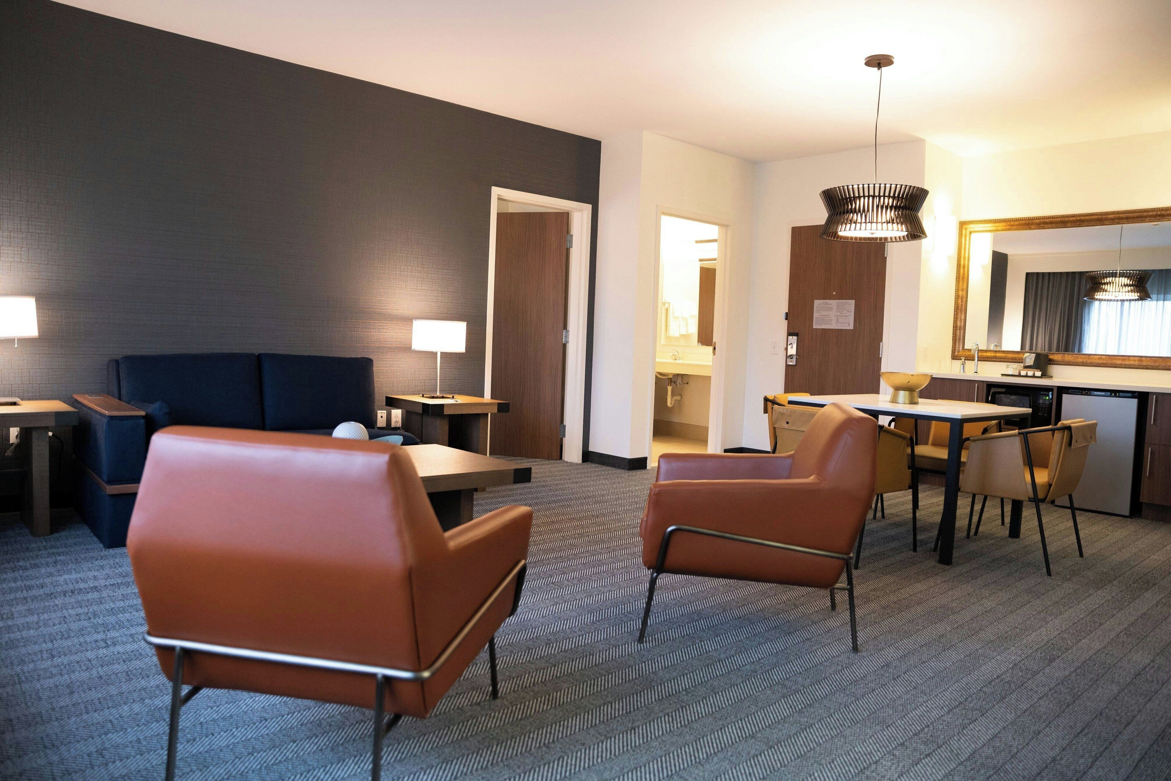 Ein Hotelzimmer mit dunkel gestrichener Wand, einem großen Spiegel, einem Schreibtisch mit Stuhl, einem Sofa und einem kleinen Tisch mit Lampe.