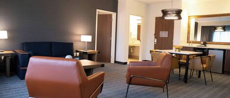 Ein Hotelzimmer mit dunkel gestrichener Wand, einem großen Spiegel, einem Schreibtisch mit Stuhl, einem Sofa und einem kleinen Tisch mit Lampe.