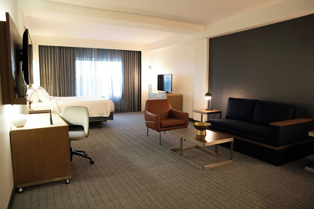 Hotellihuone, jossa on sänky, kirjoituspöytä lampun kanssa, ruskea nojatuoli, musta sohva ja jalustalla oleva televisio.