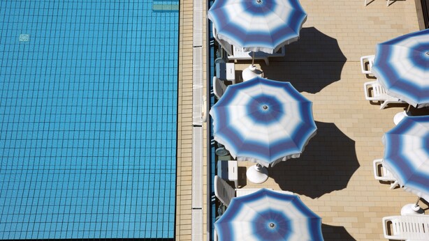 A swimming pool with tiled flooring and several lounge chairs and umbrellas arranged around it. - Hotel Antares (Cervia)