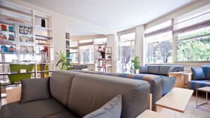A modern living room with a sectional sofa, a green chair, and a wooden coffee table. There are bookshelves and a plant in the room. - Hotel Antares (Cervia)