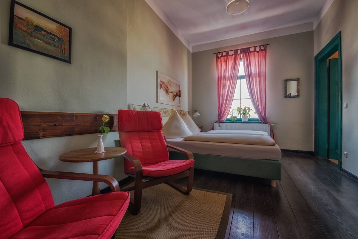 Une pièce avec un lit, un fauteuil rouge, une petite table sur laquelle se trouve un vase, et une fenêtre avec des rideaux.
