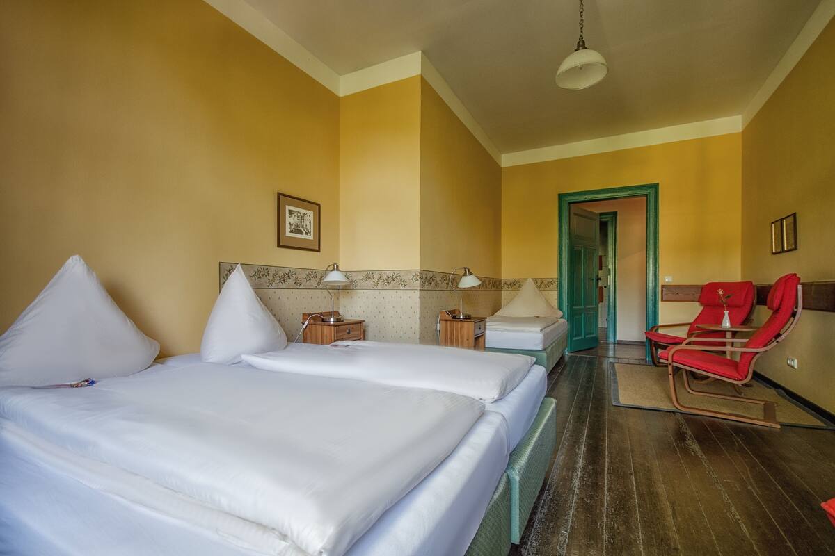 Une chambre d’hôtel avec deux lits, un fauteuil à bascule rouge et une porte verte donnant sur une autre pièce.