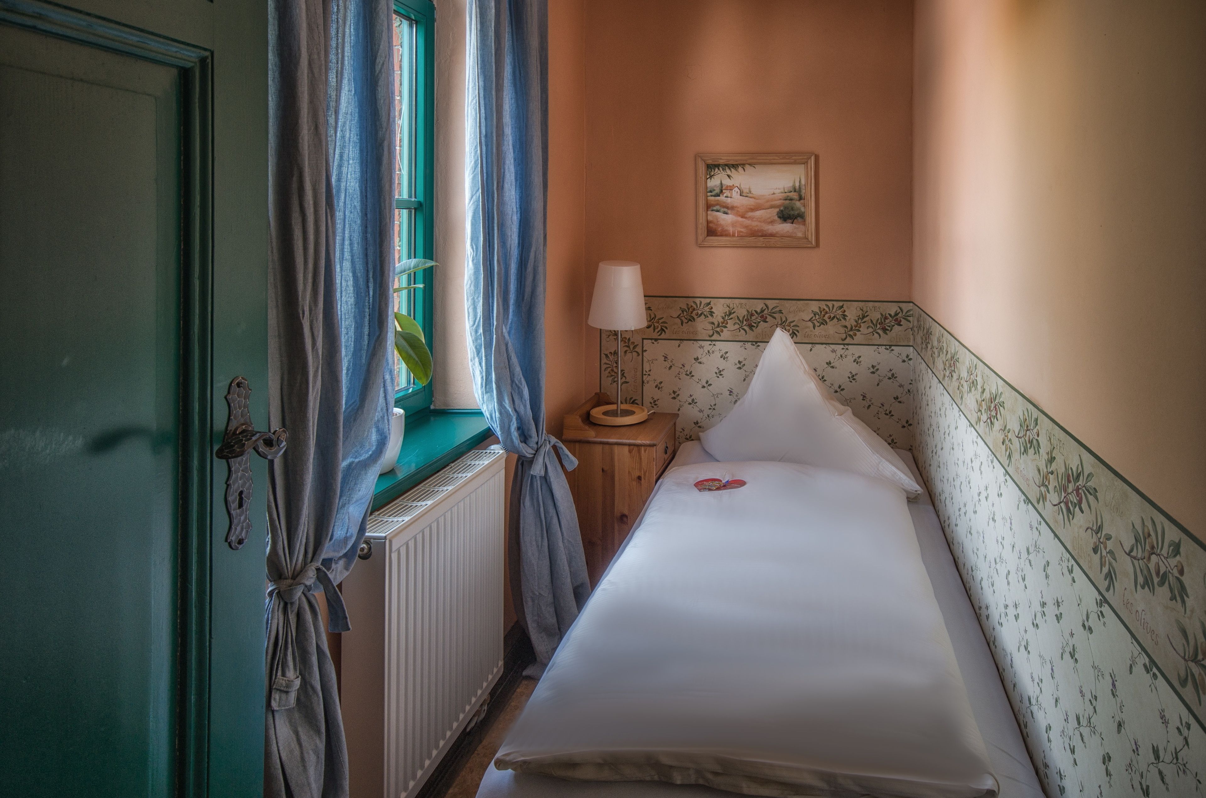 A bedroom with a bed, a nightstand, a lamp, a window with curtains, and a painting on the wall.