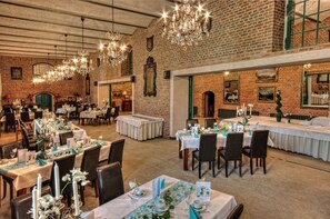 Een ruime eetruimte met meerdere tafels gedekt voor een formeel evenement, met kroonluchters en bakstenen muren.