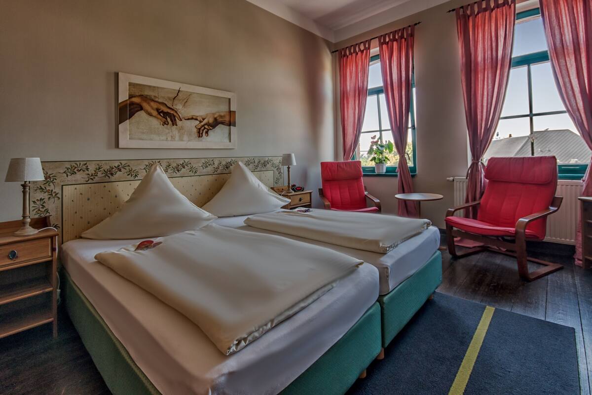 Une chambre d’hôtel avec deux lits, une chaise rouge, une petite table et un tableau accroché au mur.