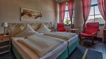 A hotel room with two beds, a red chair, a small table, and a painting on the wall.
