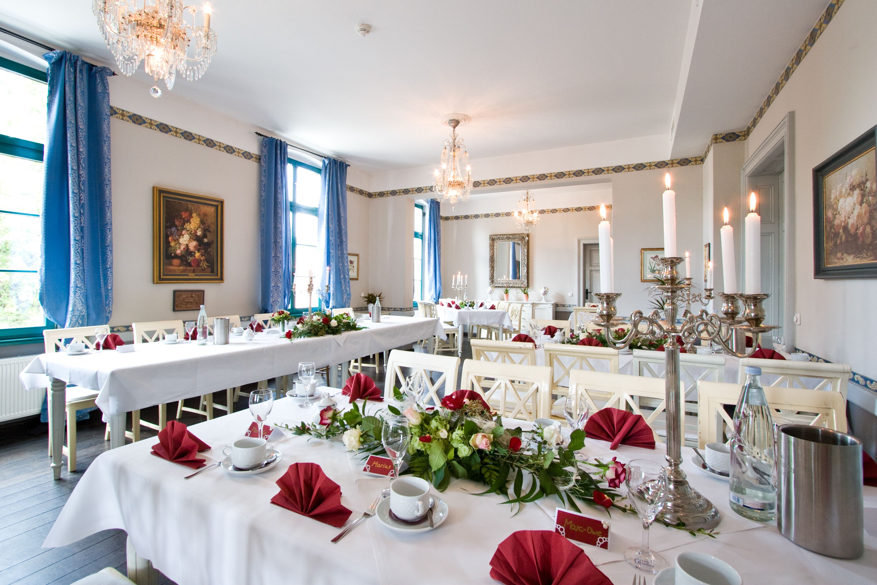 A formal dining room with long tables set for a meal, chandeliers, and large windows with blue curtains.