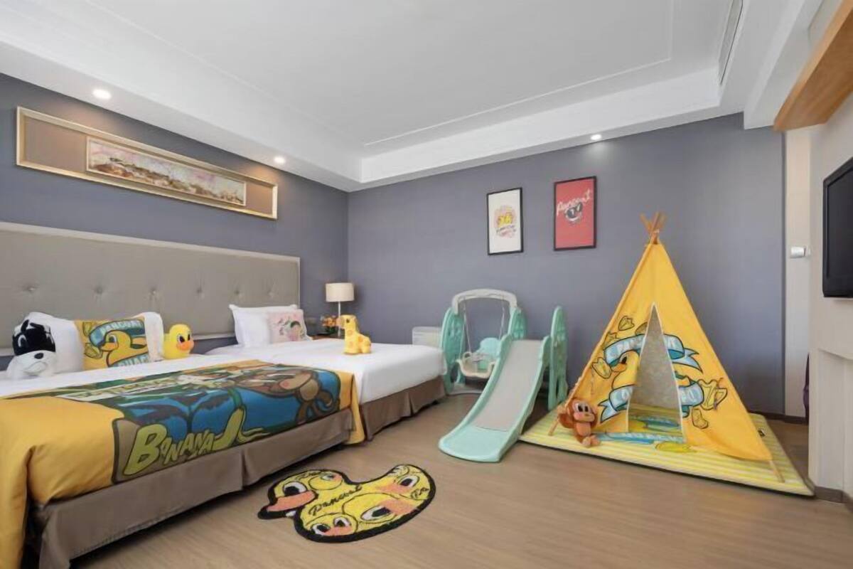 Ein Hotelzimmer mit Bett, Schreibtisch, Stuhl, Schreibtischlampe, einem an der Wand befestigten Fernseher, einem gerahmten Bild, einem gelben Zelt und einer kleinen Rutsche.