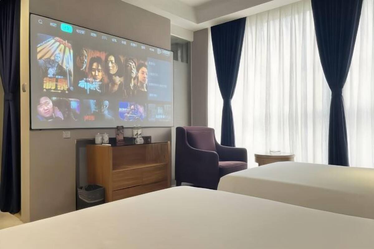 Ein Hotelzimmer mit großem Fernseher, der einen Film zeigt, einem Bett, einem Sessel und einem Fenster mit Vorhängen.