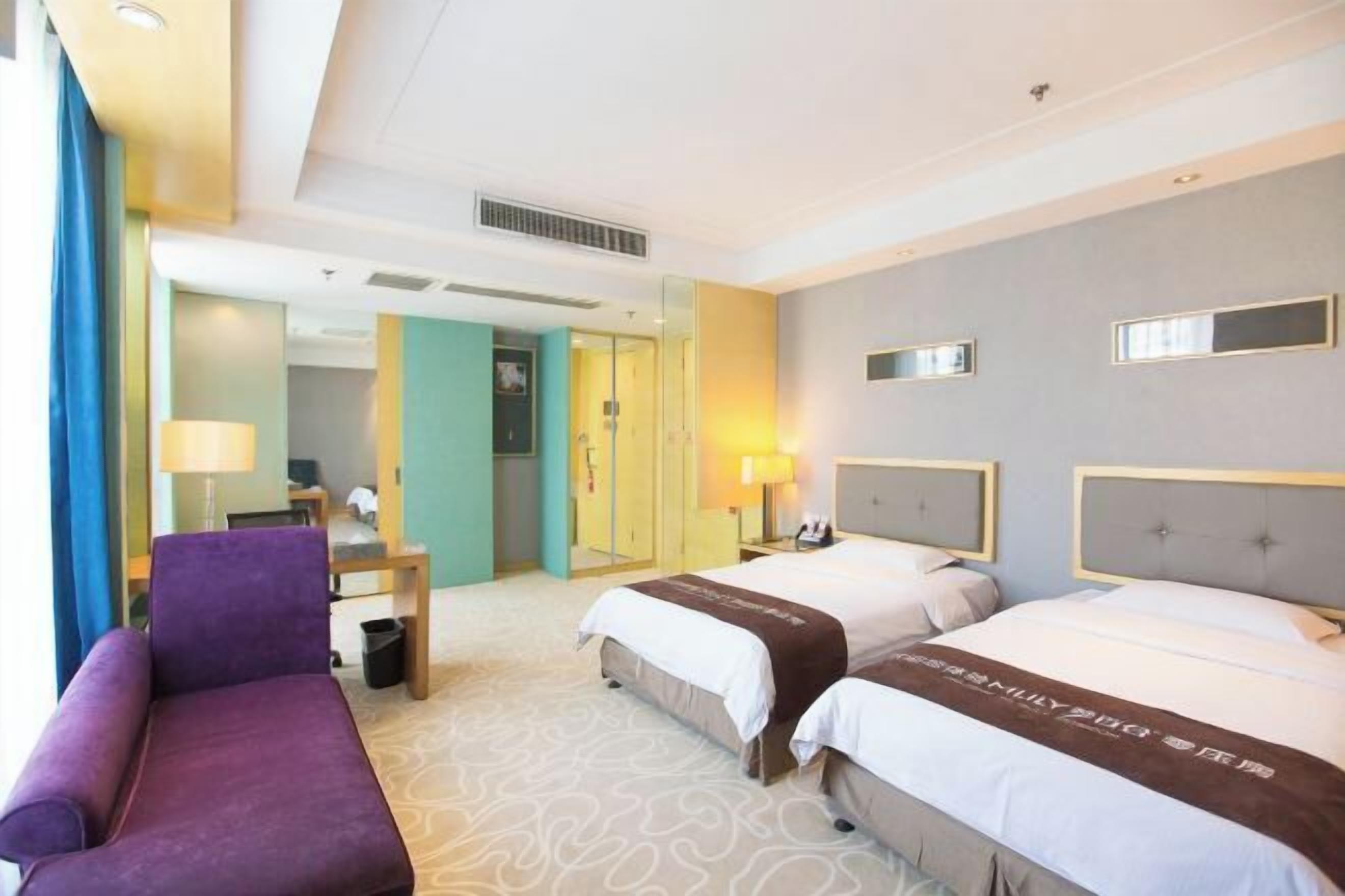 A hotel room with two beds, a purple armchair, a desk with a lamp, and a view of the hallway.
