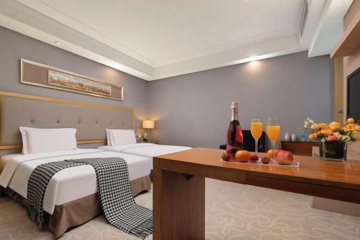 Ein Hotelzimmer mit zwei Betten, einem Holztisch mit einer Flasche, Gläsern und Obst, sowie einer Vase mit Blumen.