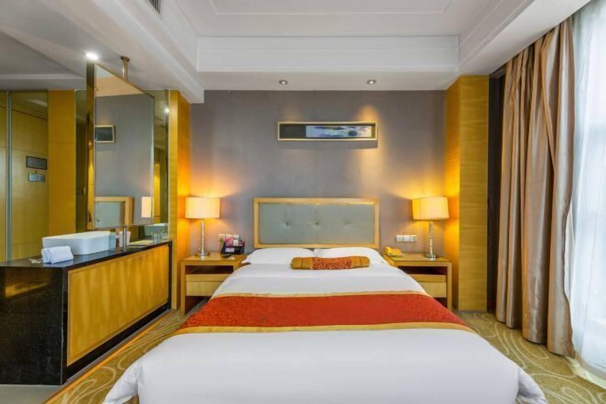 Ein Hotelzimmer mit einem großen Bett, einem Nachttisch mit Lampe, einem Spiegel und Blick ins Freie durch ein fensterbelesene Fenster.