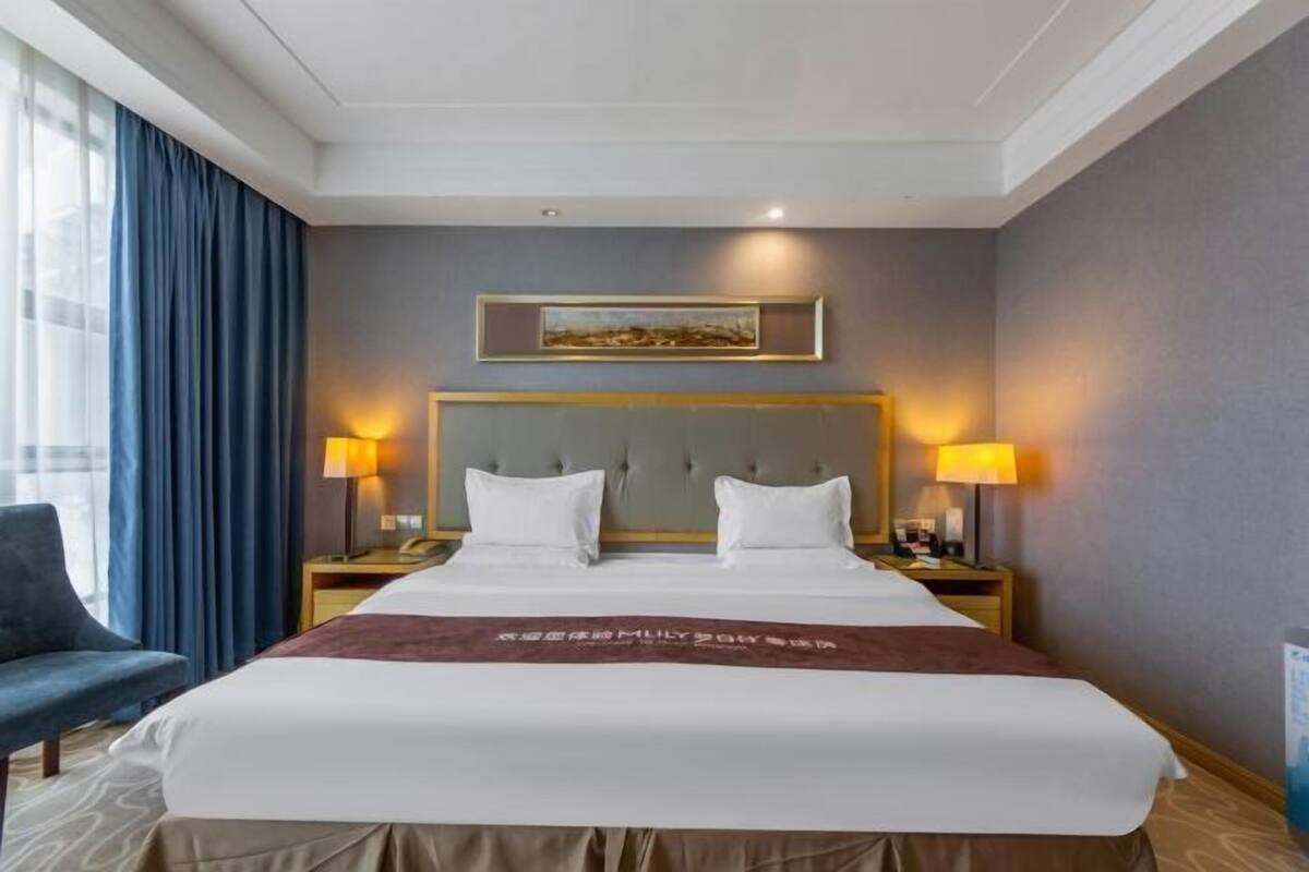 Ein Hotelzimmer mit einem großen Bett, zwei Nachttischlampen, einem Kopfteil, einem Bild an der Wand und einem blauen Vorhang.