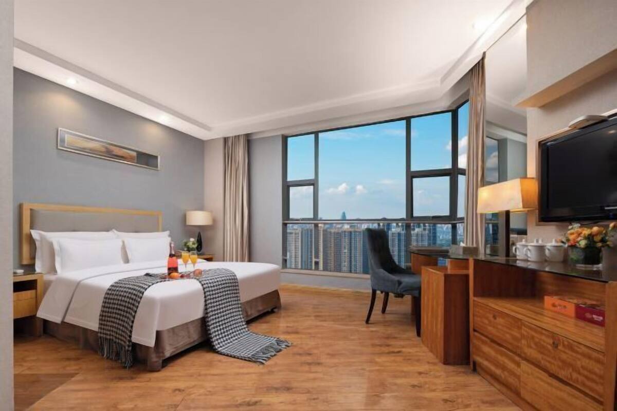 Ein Hotelzimmer mit einem Bett, einem Schreibtisch, einem Sessel, einem Fernseher und einem großen Fenster mit Blick auf die Stadt.