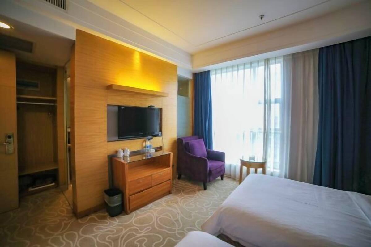 Ein Hotelzimmer mit einem Bett, einem an einer Holzplatte montierten Fernseher, einem kleinen Schreibtisch, einem violetten Sessel und einem Fenster mit Vorhängen.