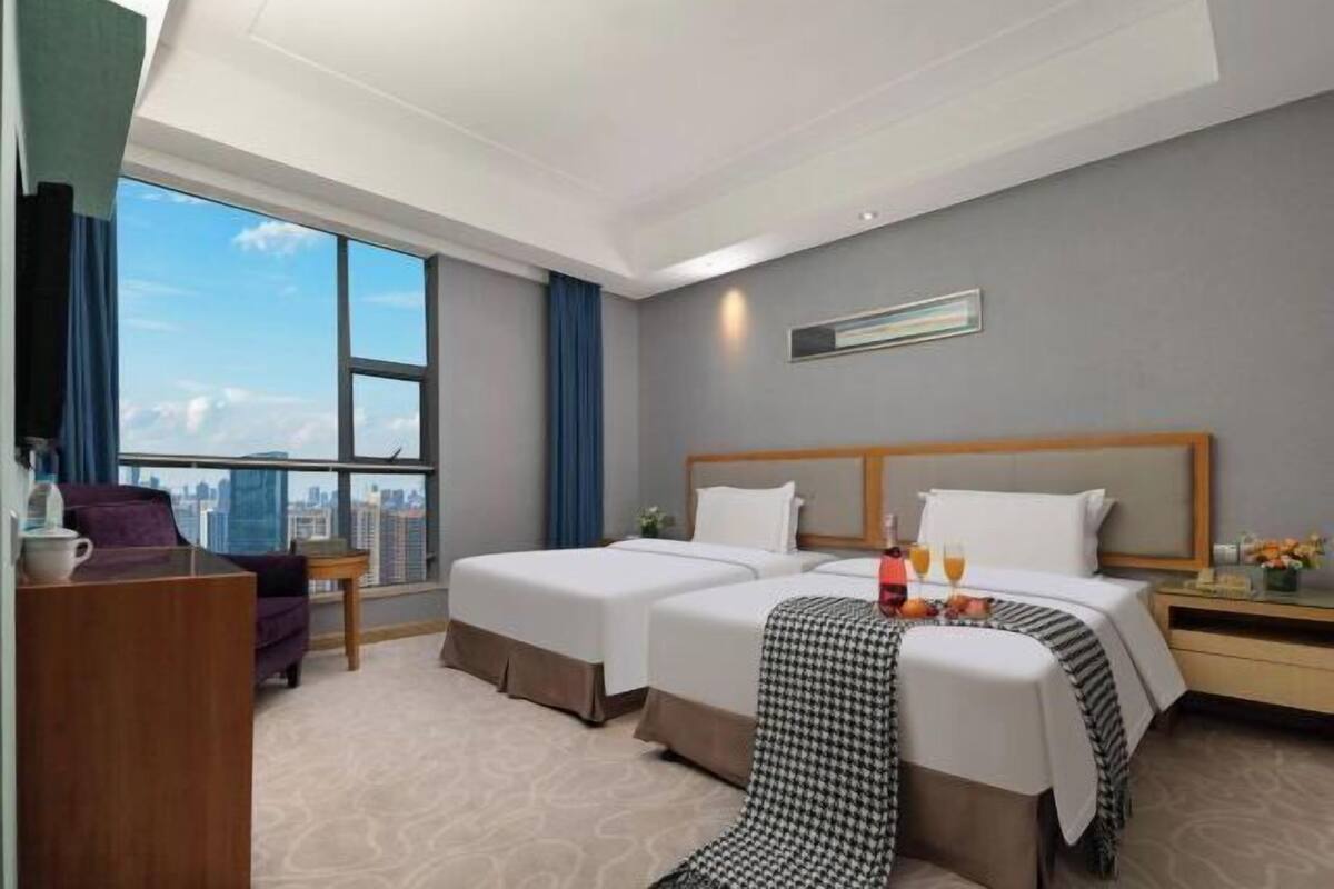 Ein Hotelzimmer mit zwei Betten, einem Schreibtisch, einem Stuhl und einem großen Fenster mit Blick auf die Stadt.