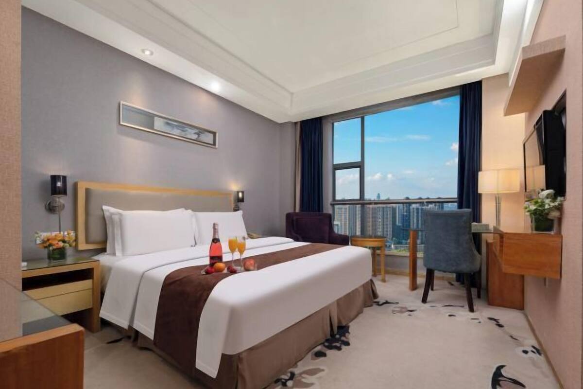 Ein Hotelzimmer mit einem großen Bett, einem Schreibtisch, einem Sessel, einem Nachttisch, einer Lampe und Blick auf die Stadt.