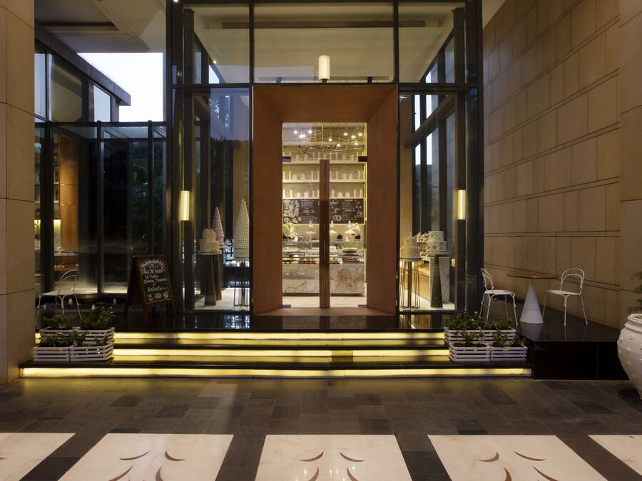 Una entrada de hotel moderna con fachada de vidrio, interior bien iluminado y un área de descanso con plantas.