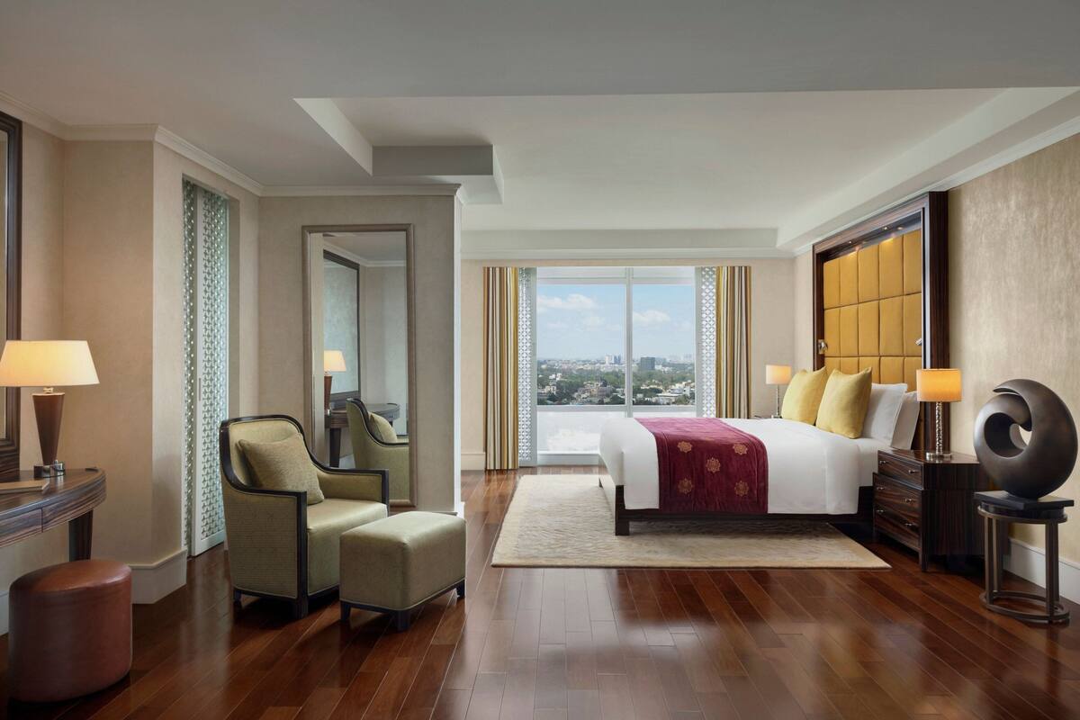 Quarto de hotel com uma cama grande, uma cadeira, uma escrivaninha e vista da cidade por amplas janelas.