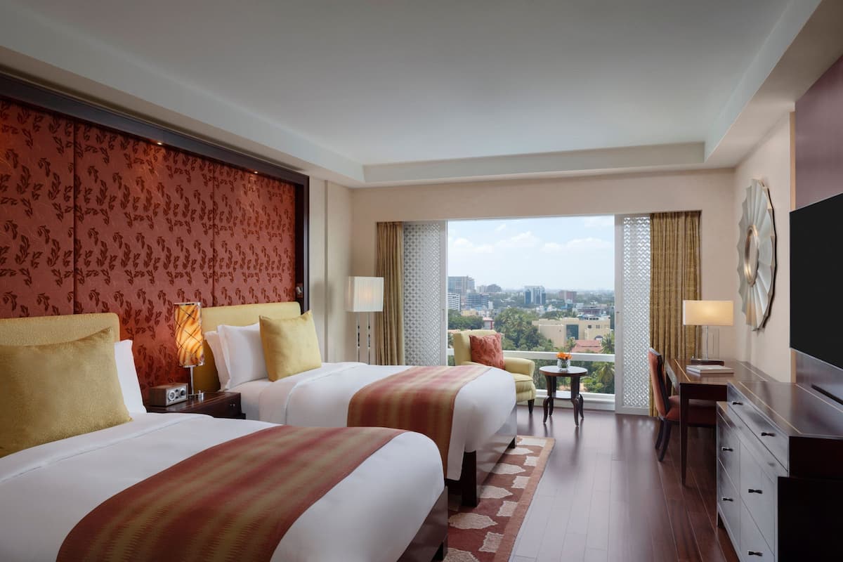 Quarto de hotel com duas camas, uma mesa, uma cadeira, televisão e varanda com vista para a cidade.