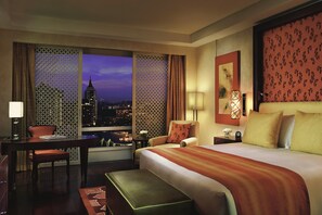 A hotel room with a large bed, a desk with a chair, a small table with a lamp, and a view of a cityscape at dusk.