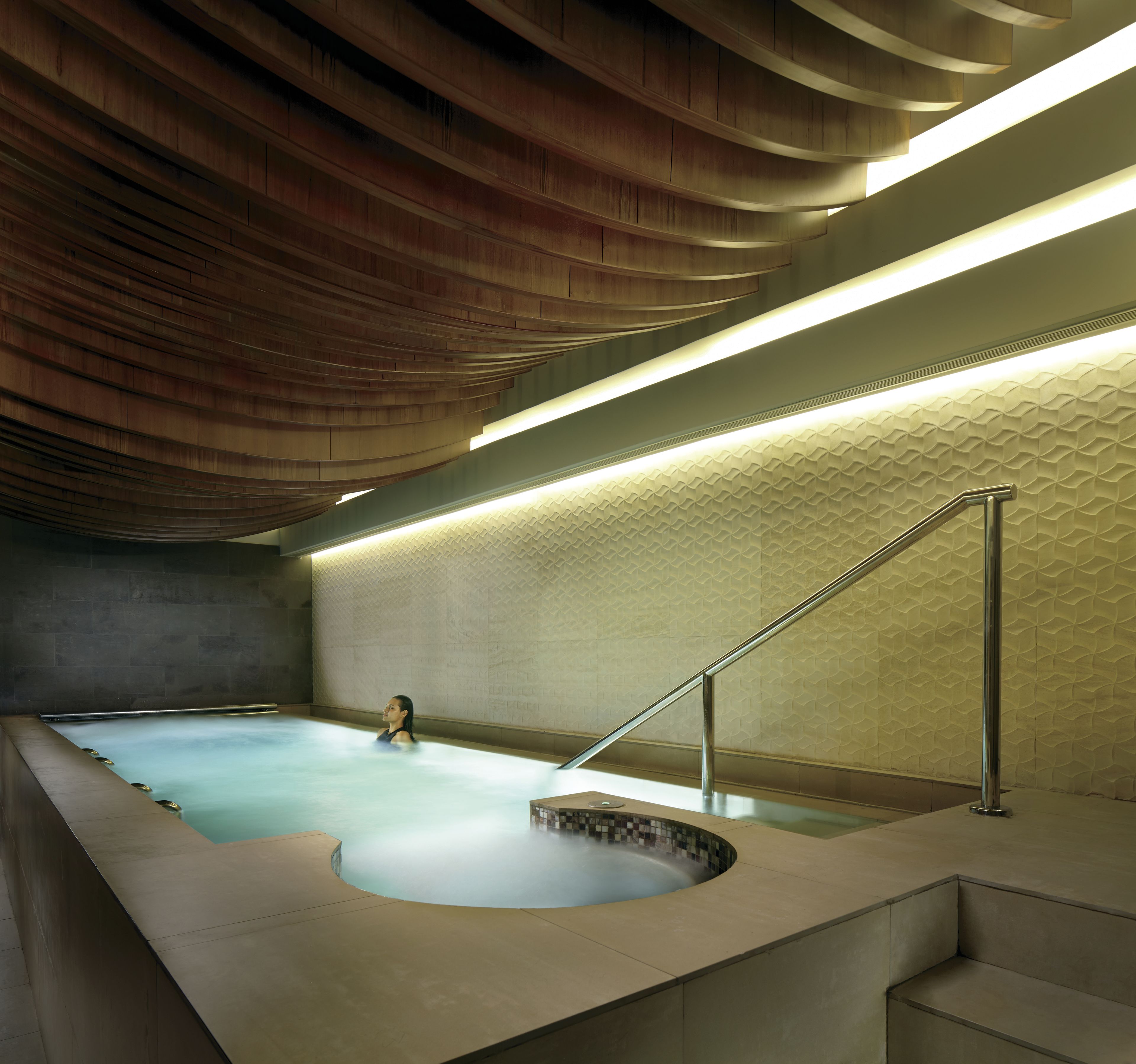 A modern indoor swimming pool with a unique wooden ceiling design and a geometric wall pattern.