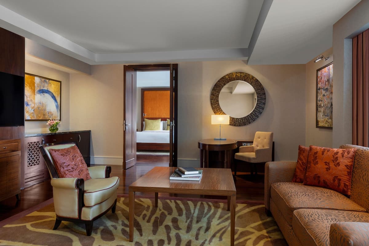 Quarto de hotel com sofá, poltrona e uma pequena mesa. Há um espelho na parede e uma porta que dá para outro quarto.