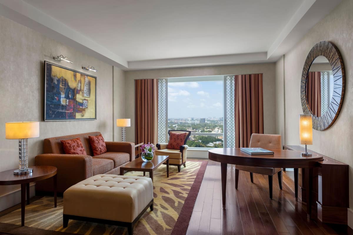 Quarto de hotel com uma janela grande, sofá, mesa de centro, mesa de jantar e um espelho decorativo.