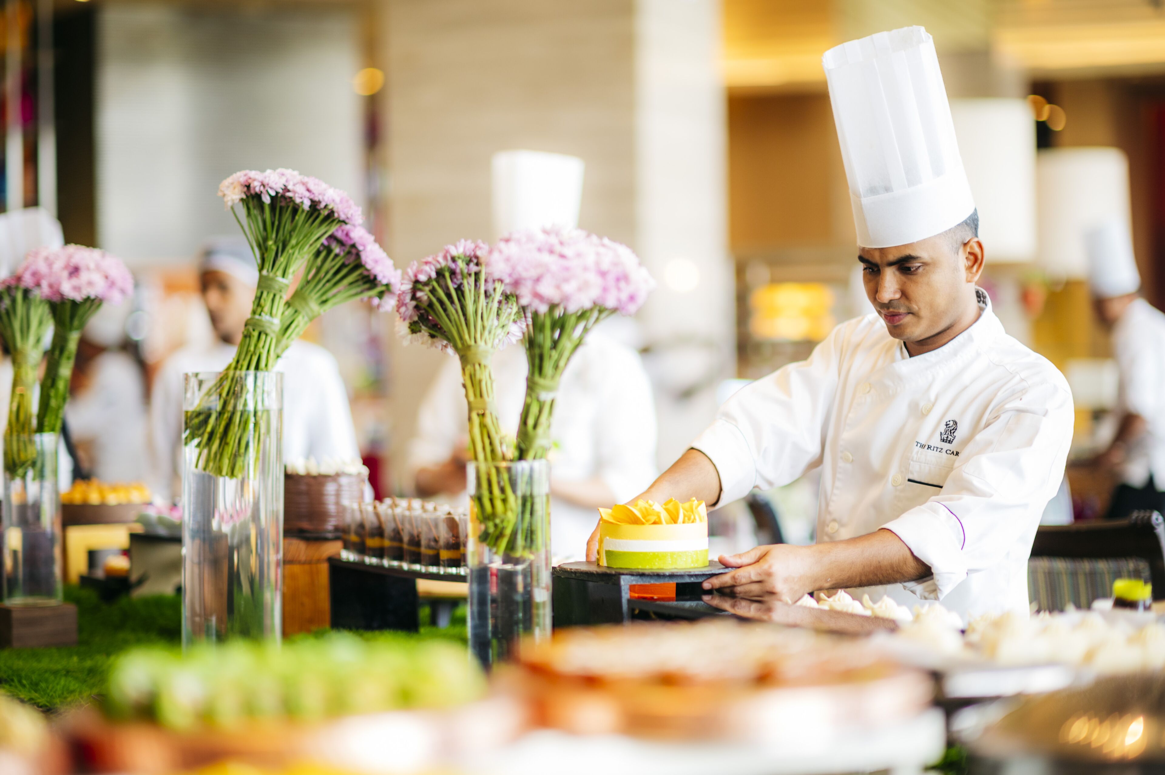 A chef in a white uniform with a tall hat, preparing food at a counter with a glass vase containing pink flowers.