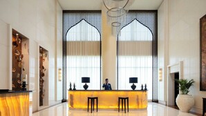 A hotel reception area with a marble counter, two armchairs, a chandelier, and a large window with a decorative screen.