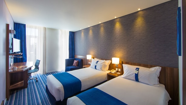 A hotel room with two beds, a desk, a chair, and a large window with curtains.