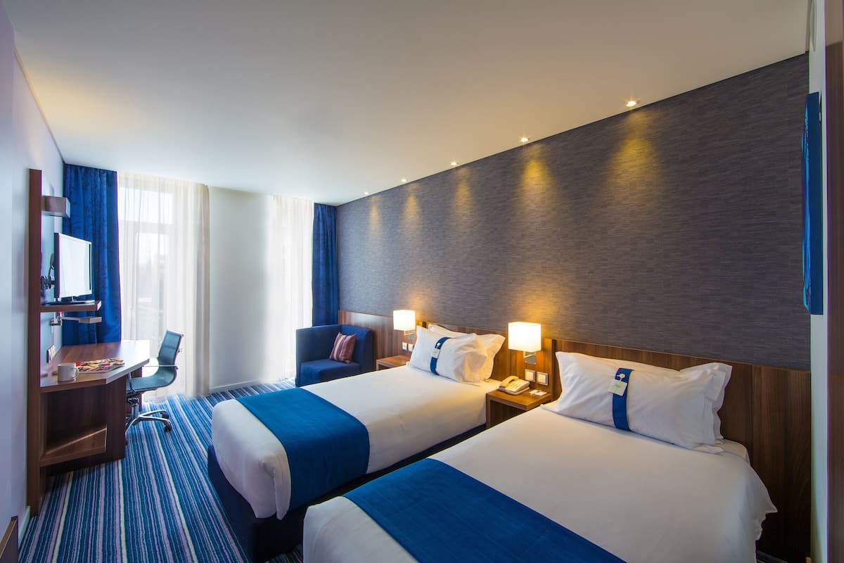 Habitación de hotel con dos camas, un escritorio, una silla y un ventanal con cortinas.