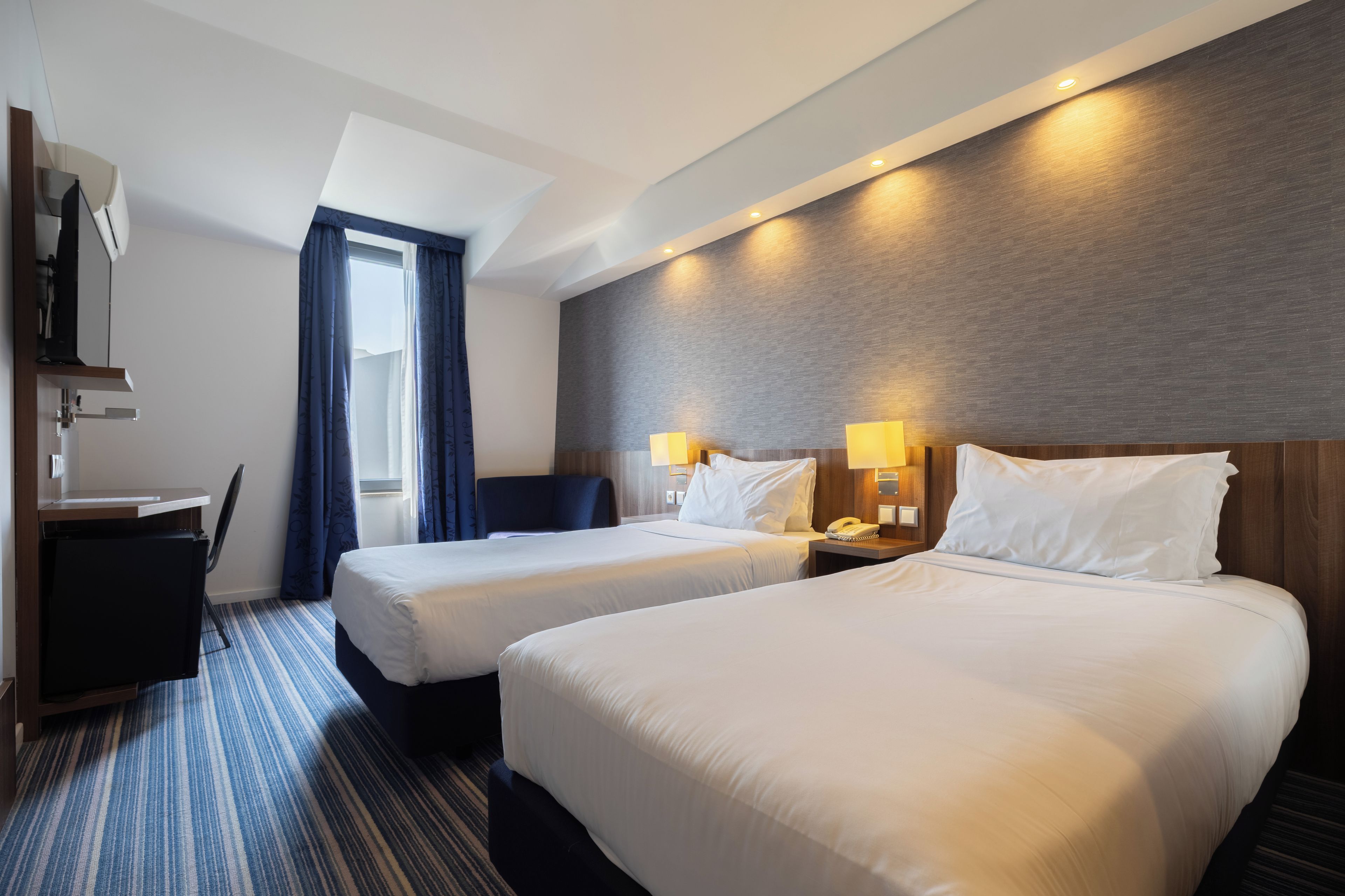 A hotel room with two beds, a desk, a chair, a television, and a window with curtains.
