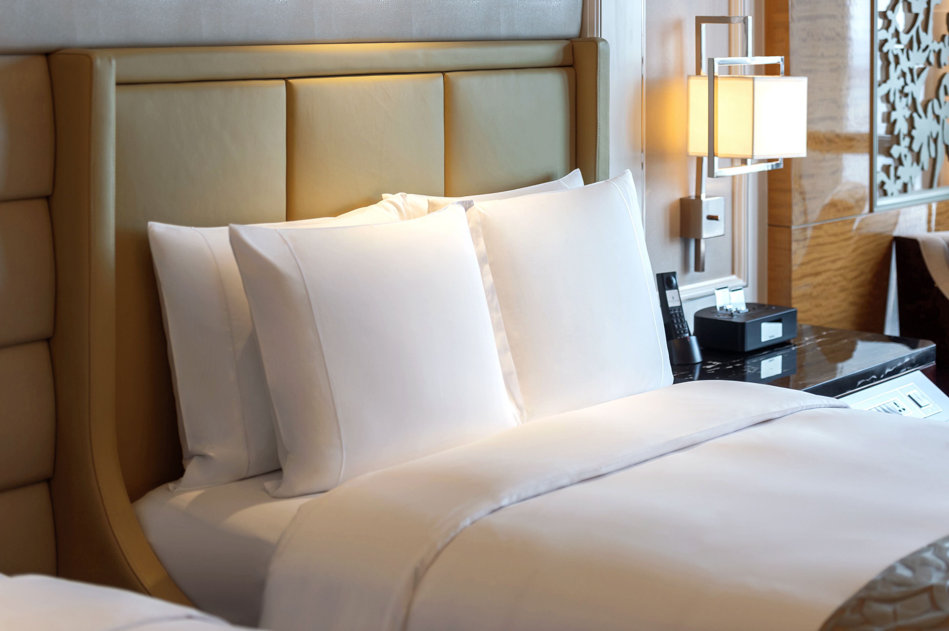 Una habitación de hotel con una cama grande, un cabecero con un panel de color claro, una mesita de noche con lámpara y un panel decorativo en la pared.