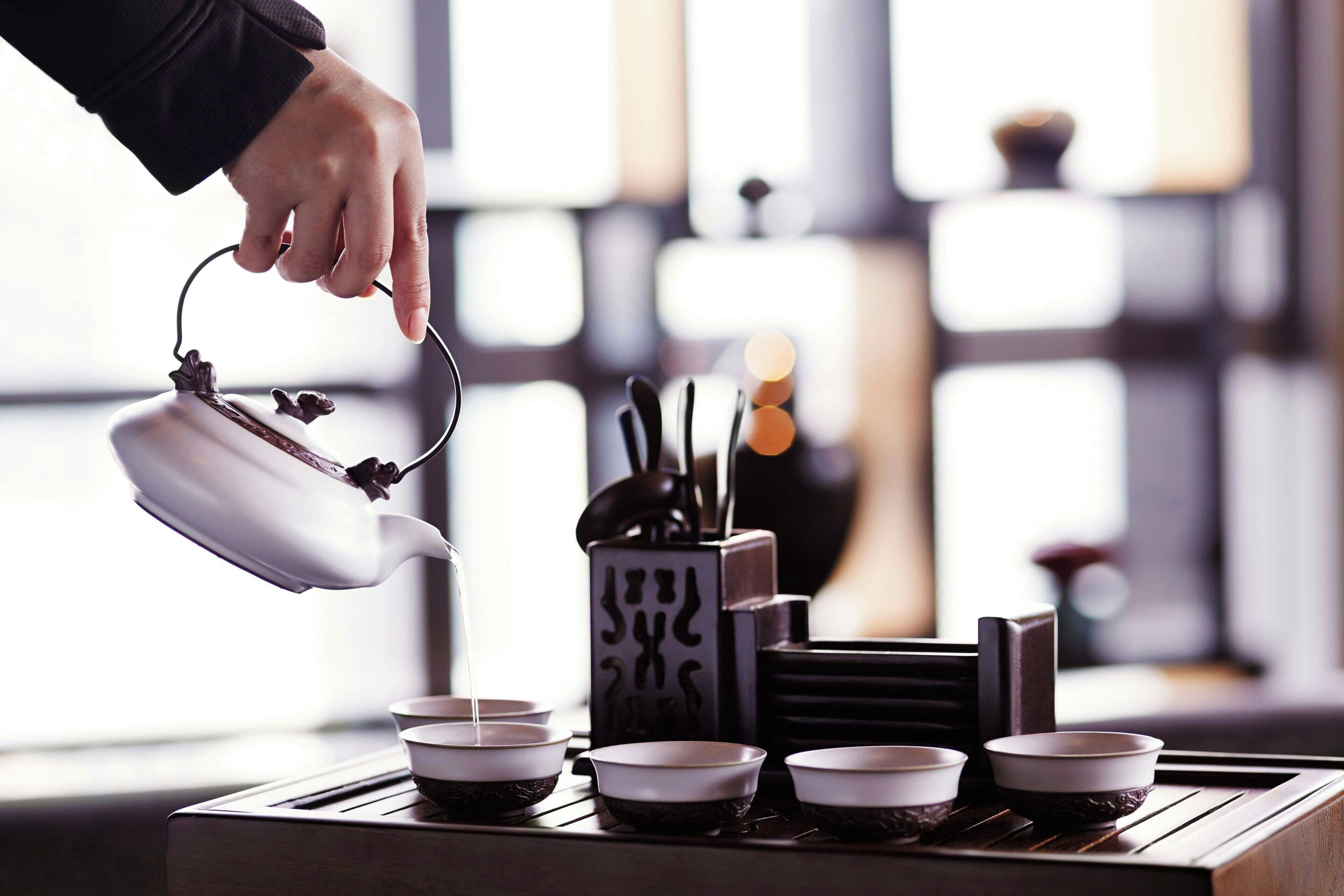 A hand pouring tea from a teapot into a cup, with several other cups and a tea set on a tray.