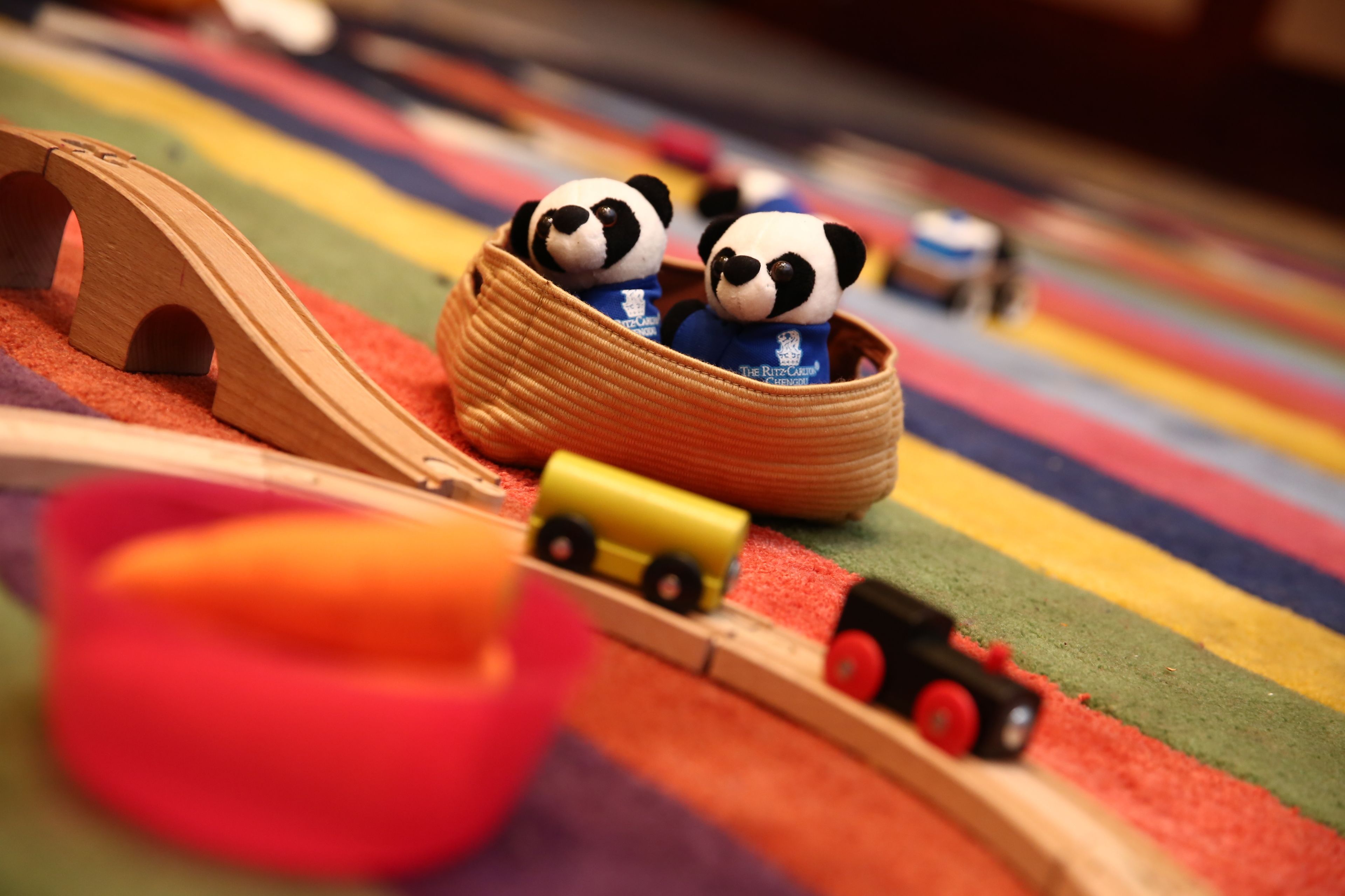 Un tren de juguete de madera con dos figuras de panda en una cesta, un vagón amarillo y un vagón rojo sobre una superficie de rayas coloridas.