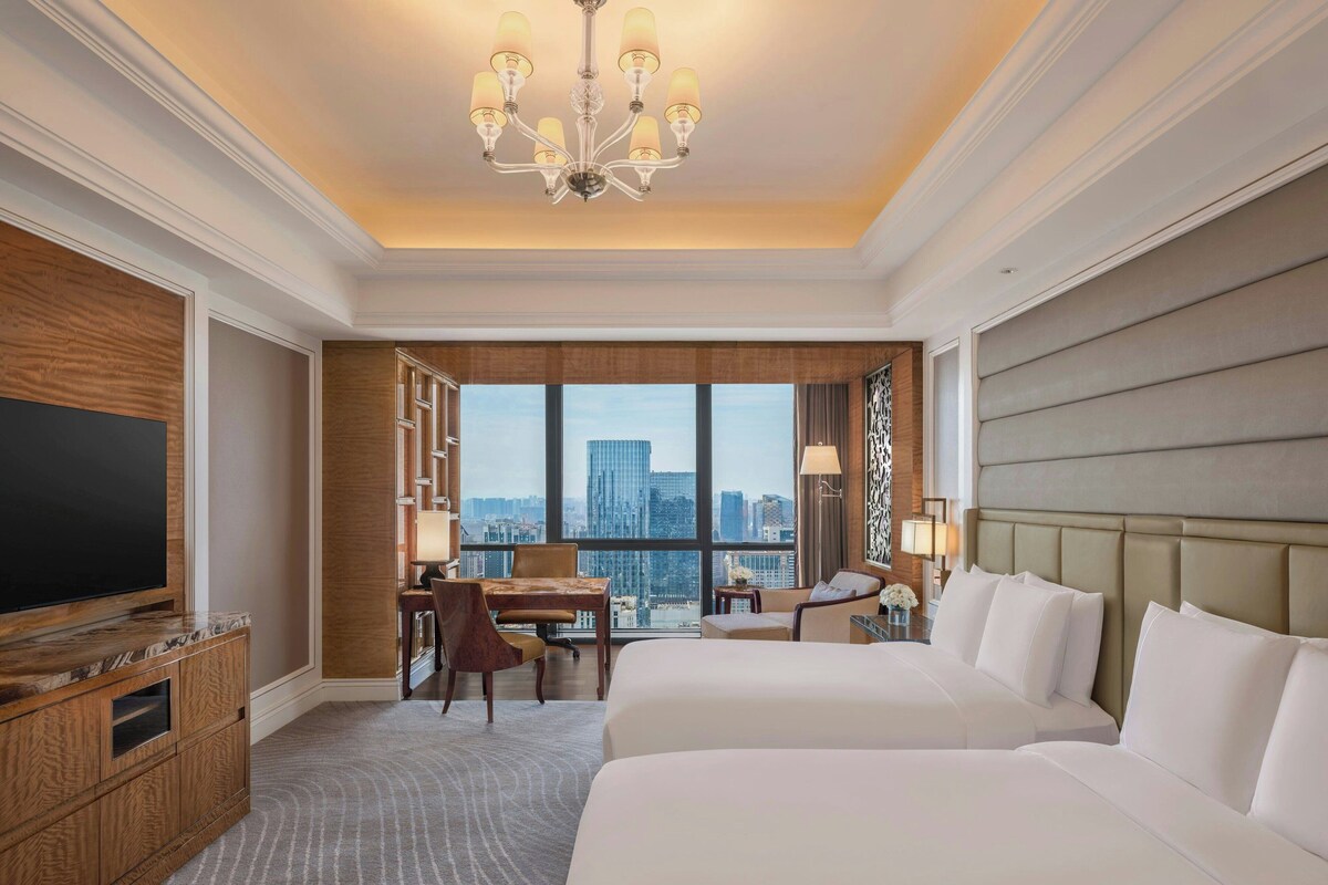 A hotel room with a large bed, a desk, a television, and a view of the cityscape.