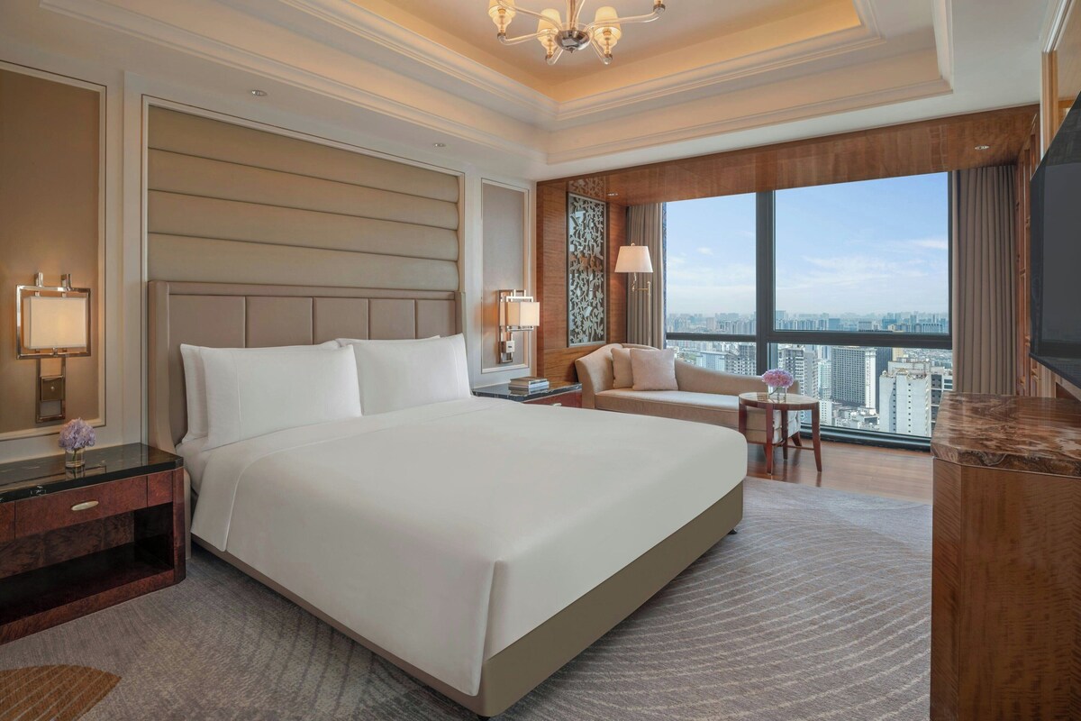 A hotel room with a large bed, a desk, a chair, a TV, a window with a city view, and a decorative wall panel.