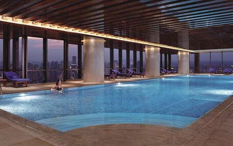 An indoor swimming pool with a city view, large pillars, and lounge chairs.