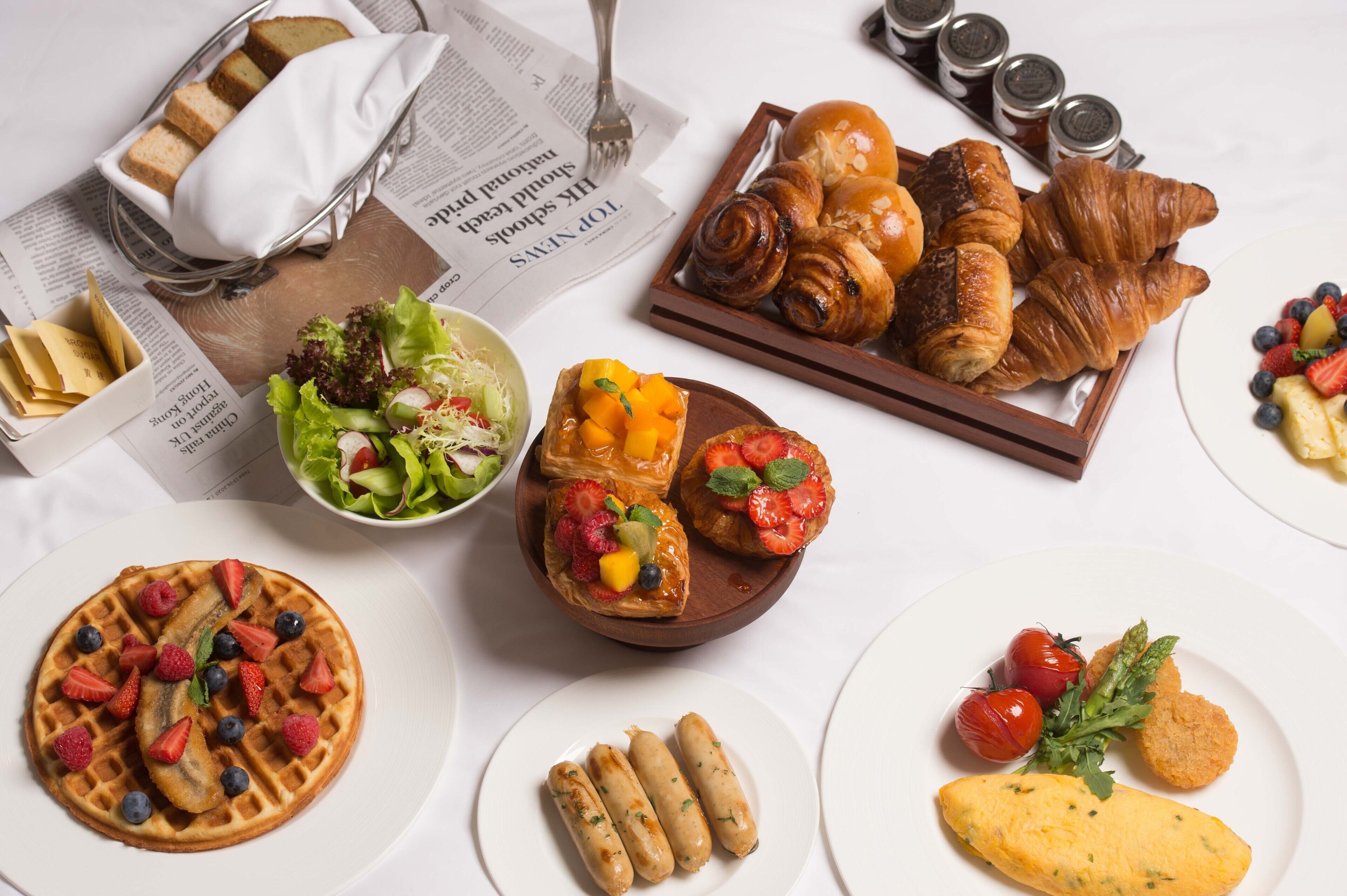 A variety of breakfast items including waffles, pastries, and a salad on a white table.
