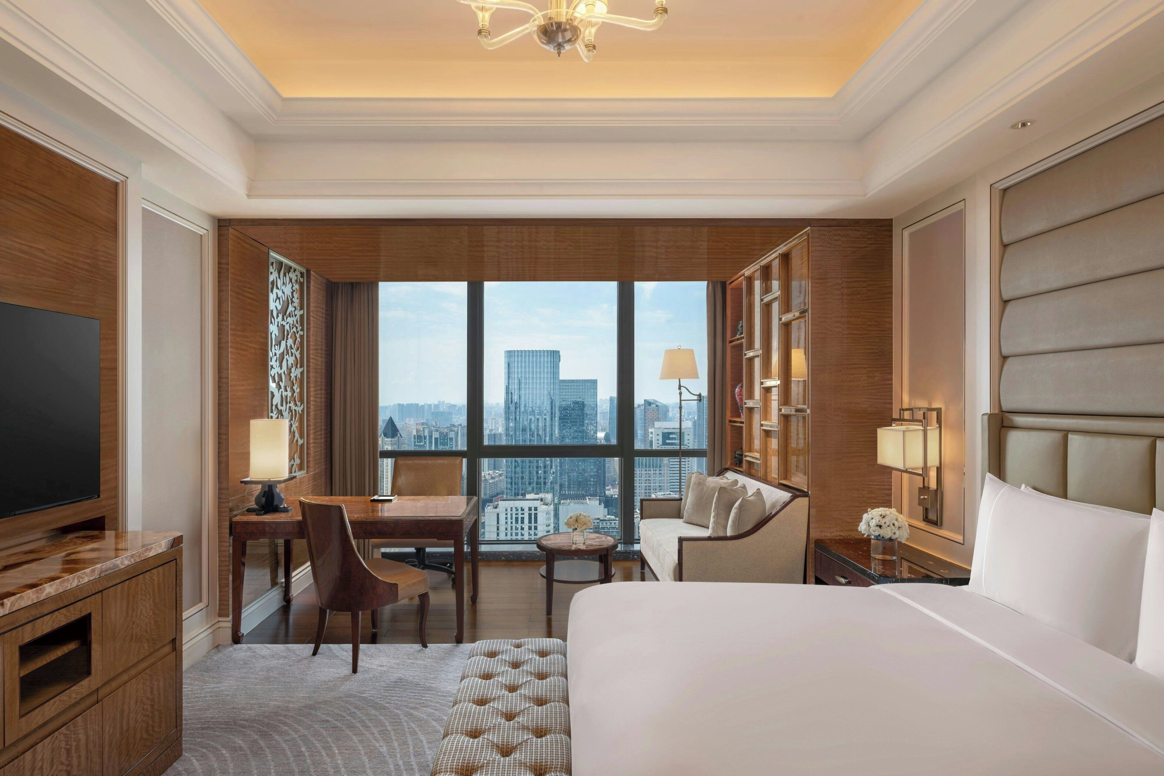 A hotel room with a large bed, a desk with a chair, a television, and a view of the cityscape through large windows.