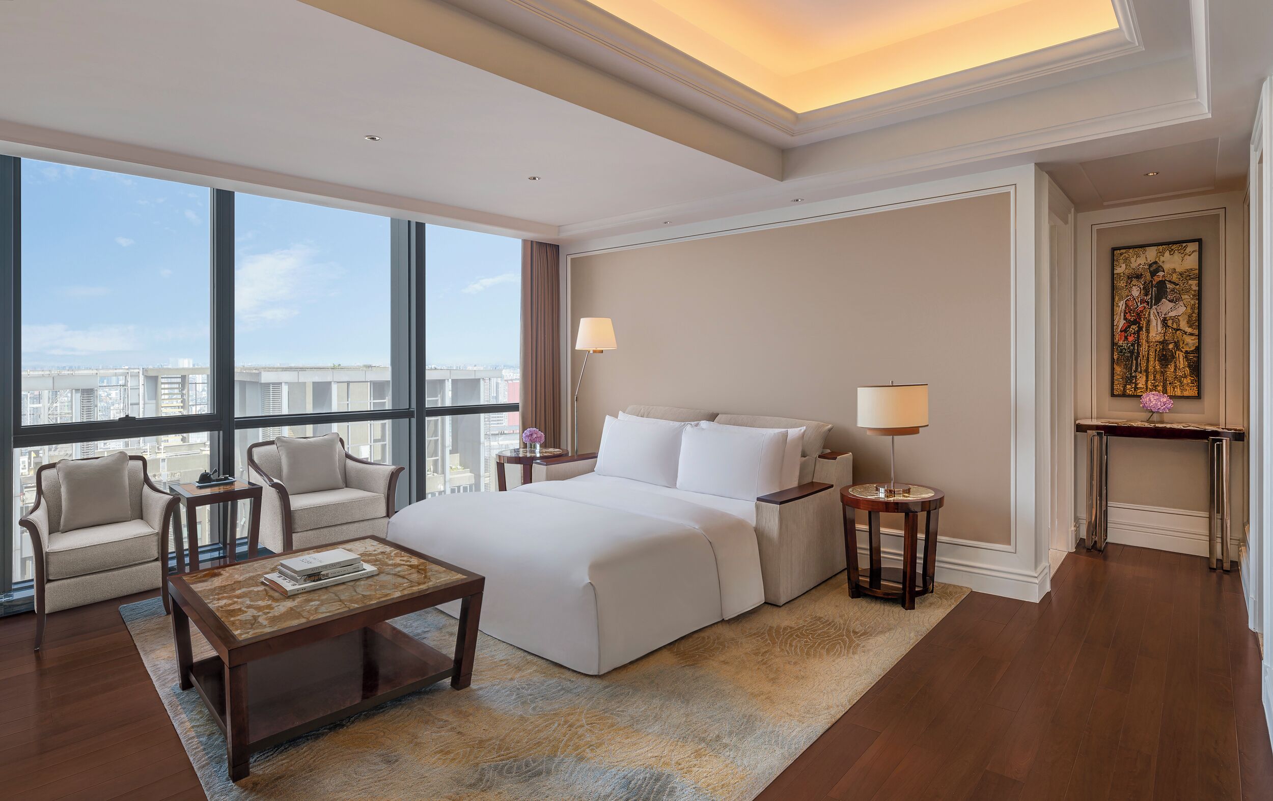A hotel room with a large bed, a sitting area with a chair and a table, a small side table, and a painting on the wall.