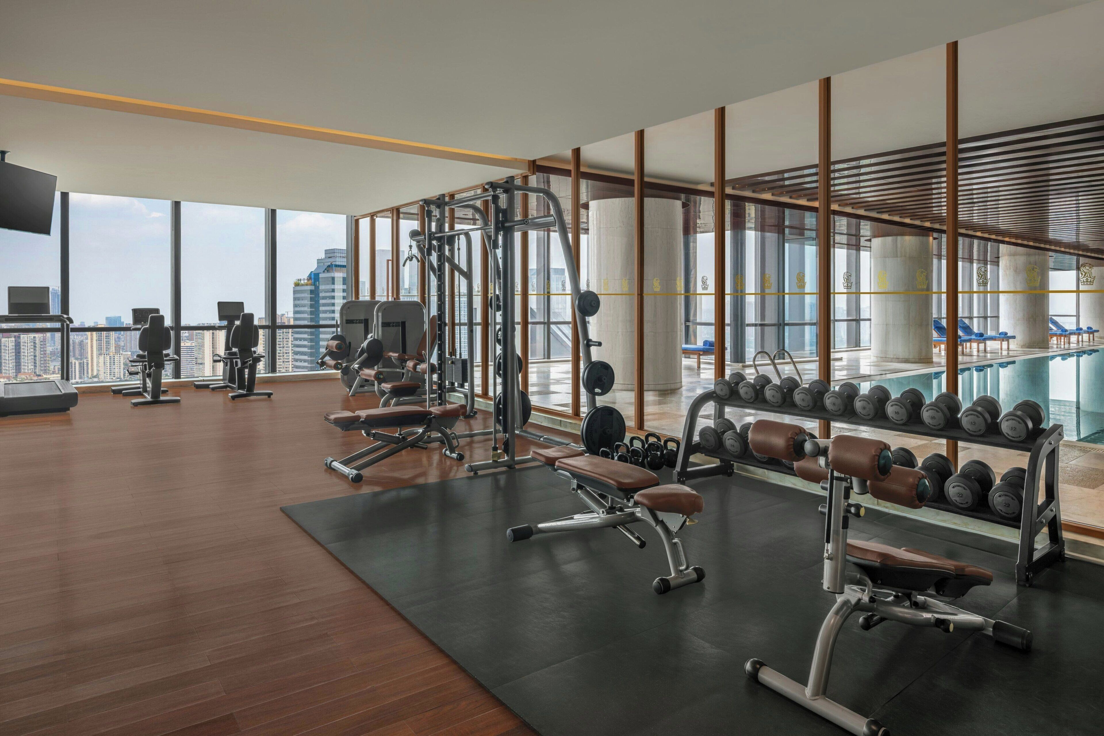 Une salle de sport moderne équipée de divers appareils d’exercice, dotée d’une grande fenêtre offrant une vue sur la ville et d’un espace piscine visible à travers la vitre.