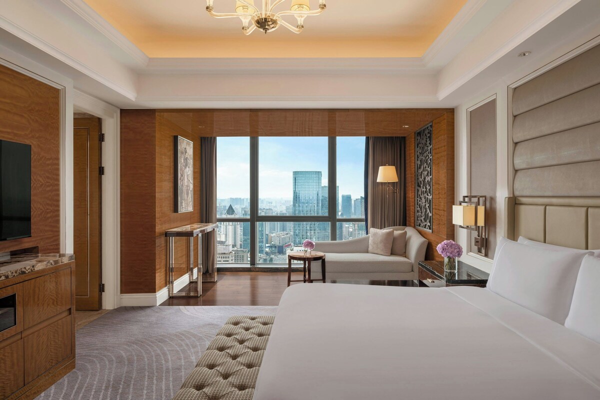 A hotel room with a large bed, a television, a desk, and a view of the cityscape.
