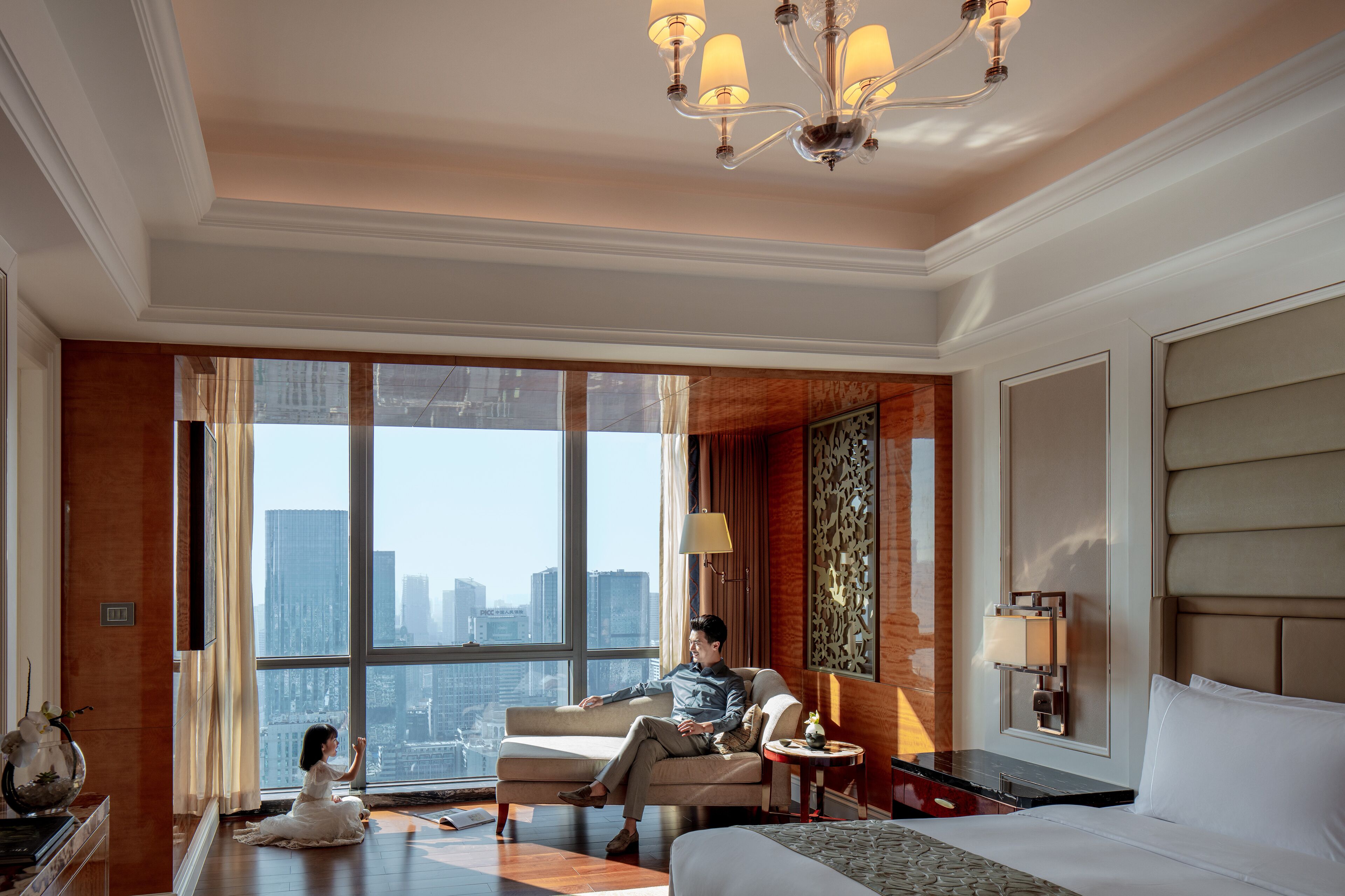 A spacious hotel room with a large window offering a city view, a comfortable sofa, a bed, and a small table with a lamp.